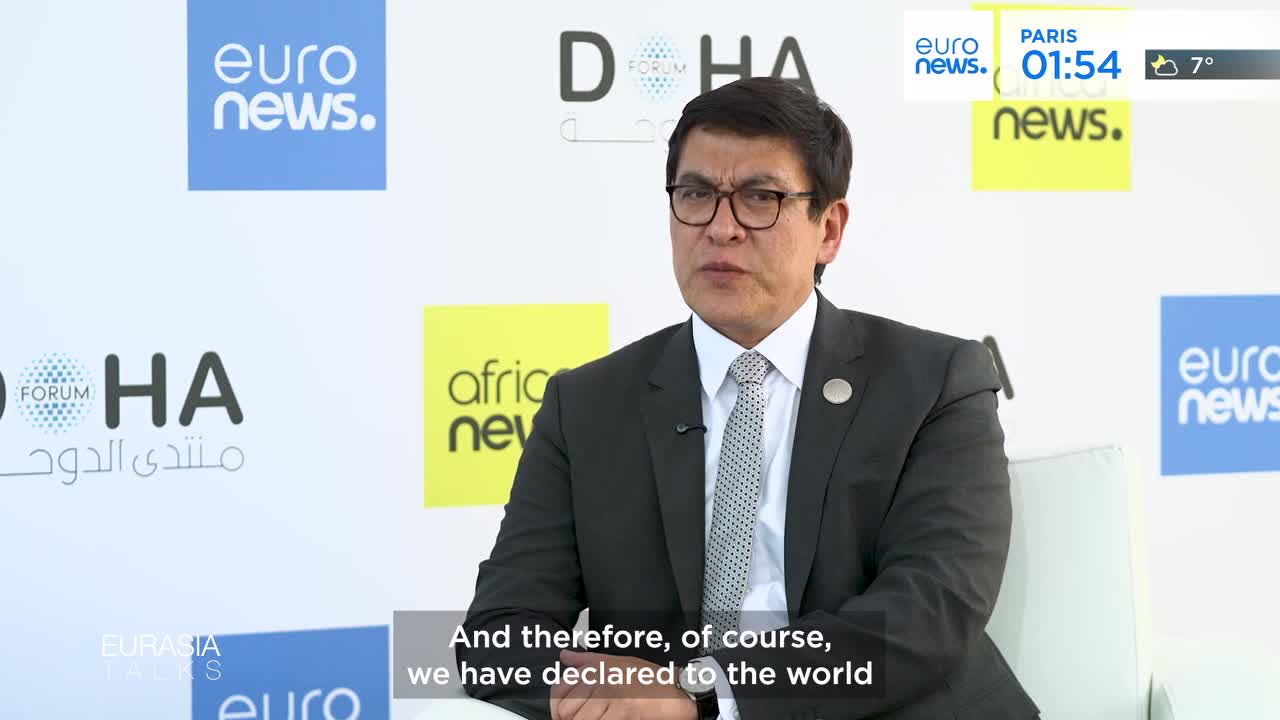 A man in a suit speaks into a microphone, his glasses reflecting the light. Behind him, a screen displays "Doha Forum" and "Euronews."