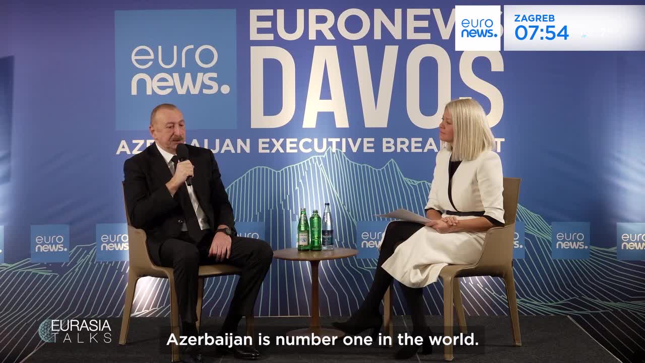 A man in a dark suit speaks into a microphone, seated next to a woman in a white jacket. They are positioned in front of a large blue backdrop with "EURONEWS DAVOS" printed across it.