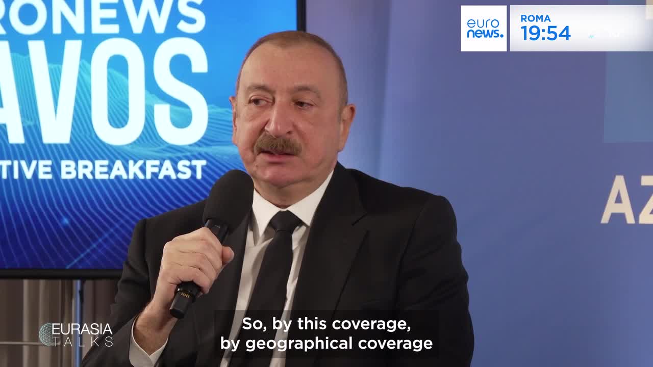 A man in a suit speaks into a microphone, his hand resting on it. Behind him, a screen displays "EURONEWS DAVOS" and "EURASIA TALKS." A clock in the corner shows 19:54.
