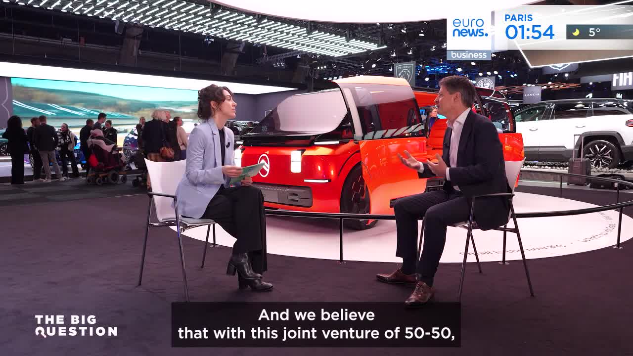 A woman in a light blue blazer sits opposite a man in a dark suit, both facing a bright orange concept car. Behind them, a crowd mills around other vehicles at what looks like a Paris auto show.