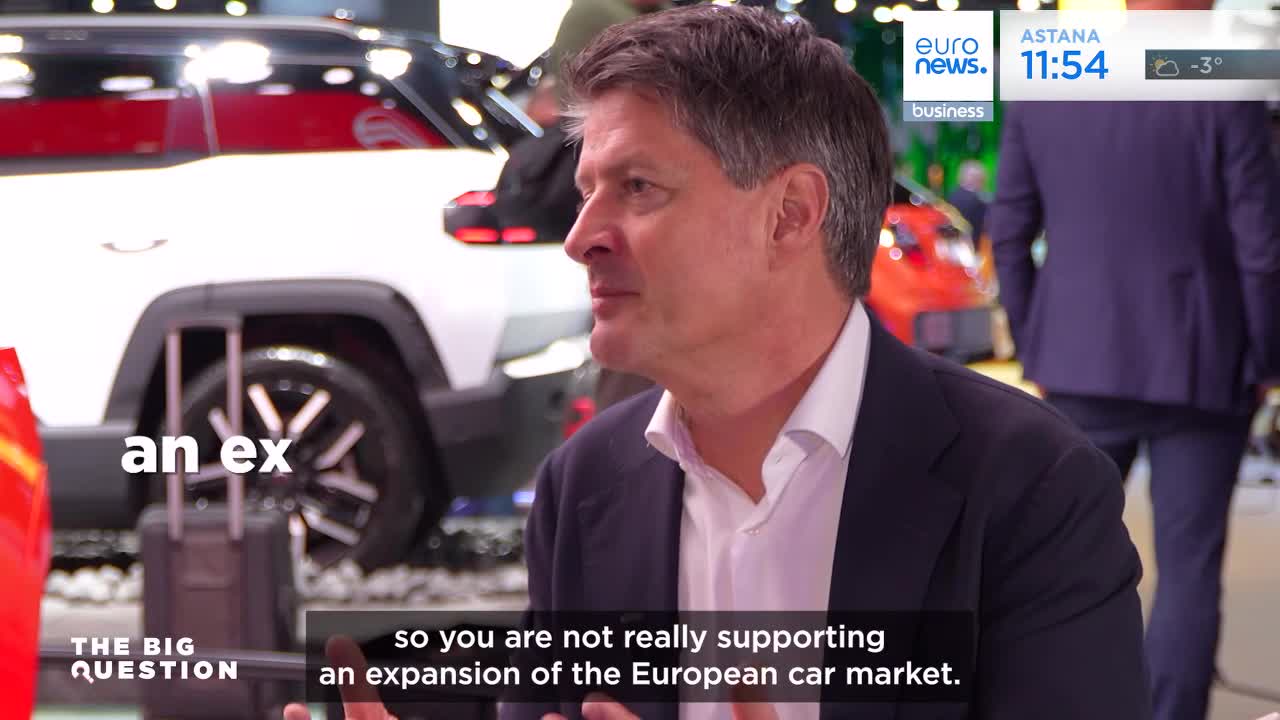 A man in a dark suit and white shirt speaks, his profile turned to the side. Behind him, a white car with black accents is on display, and a red car is partially visible.