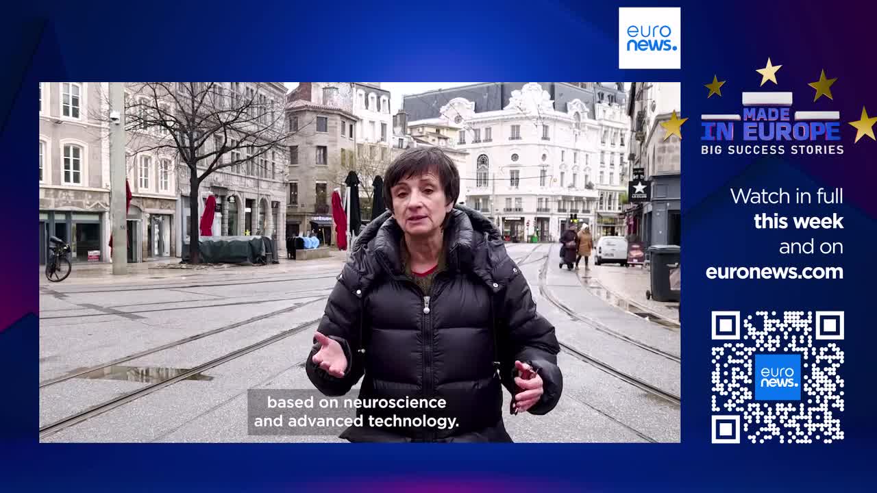 A woman speaks directly to the camera on a street in France, with tram tracks running through the cobblestones. In the background, a few people walk past shops lining the wide avenue.