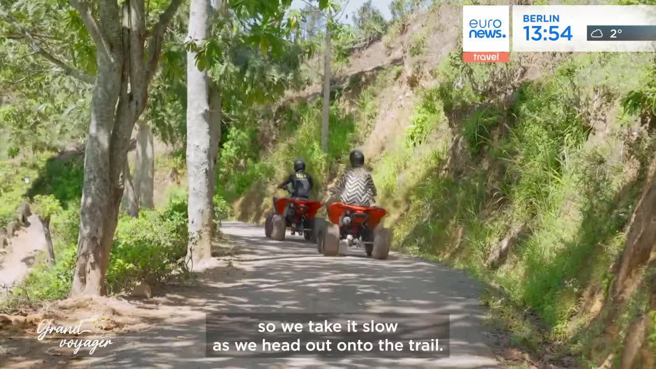 Two red ATVs, each with a rider, rumble down a narrow dirt path lined by lush green foliage. The lead rider wears a patterned shirt, while the one behind them is in a dark jacket.