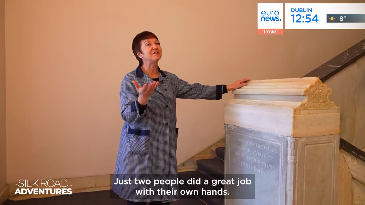 A woman in a patterned coat gestures with her hands, speaking animatedly. She rests one hand on a carved stone structure, its surface covered in faint inscriptions. A woman in a patterned coat gestures with her hands, speaking animatedly. She rests one hand on a carved stone structure, its surface covered in faint inscriptions.