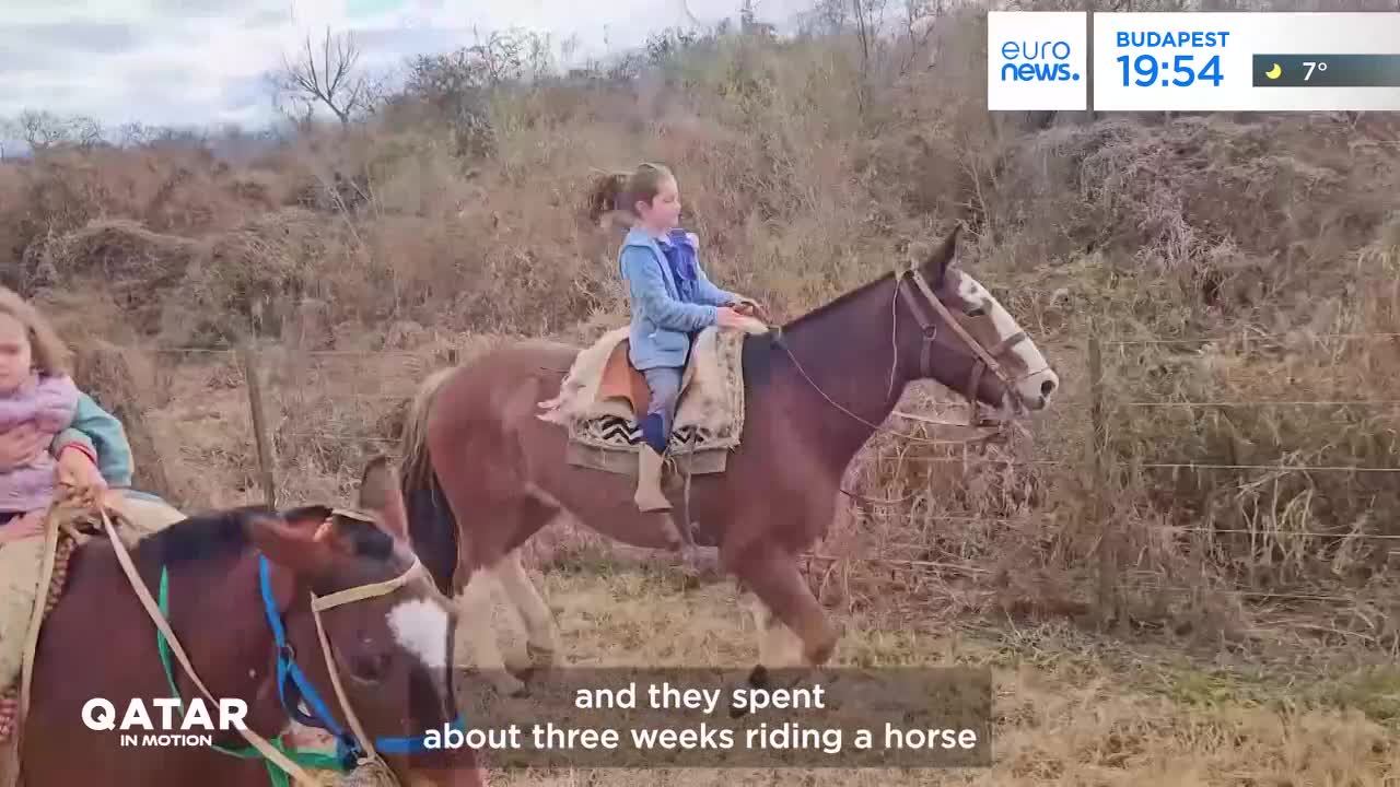A young girl in a blue jacket rides a brown horse through dry brush. Another child on a horse follows closely behind. A young girl in a blue jacket rides a brown horse through dry brush. Another child on a horse follows closely behind.