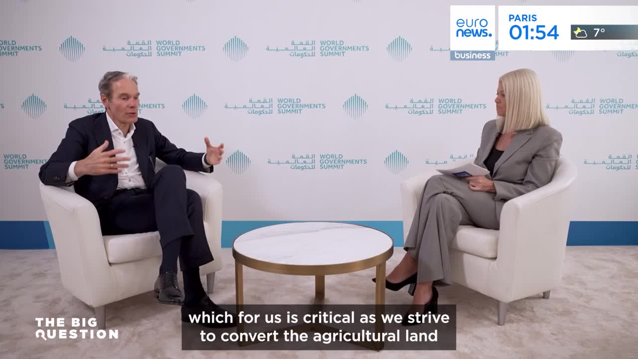 A man in a suit gestures with his hands as he speaks, facing a woman in a grey suit who holds papers. The backdrop behind them is emblazoned with "World Governments Summit." A man in a suit gestures with his hands as he speaks, facing a woman in a grey suit who holds papers. The backdrop behind them is emblazoned with "World Governments Summit."