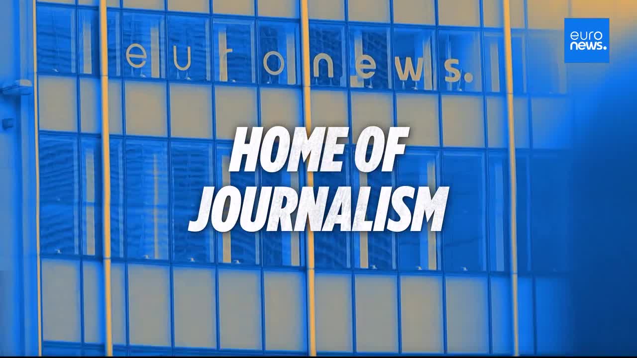 The glass facade of the Euronews building in France displays the words "HOME OF JOURNALISM" in large white letters. Sunlight catches the windows, reflecting the blue and yellow hues of the Euronews logo.