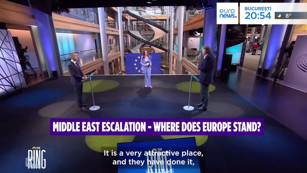 A man in a dark suit stands at a podium, facing left, while a woman in a light blue suit stands center, holding a microphone. Another man, dressed in a blazer, stands at a podium to the right, facing the woman. The backdrop features a large European Union flag and a modern, multi-story interior with a prominent spiral staircase. A graphic at the bottom reads "MIDDLE EAST ESCALATION - WHERE DOES EUROPE STAND?"