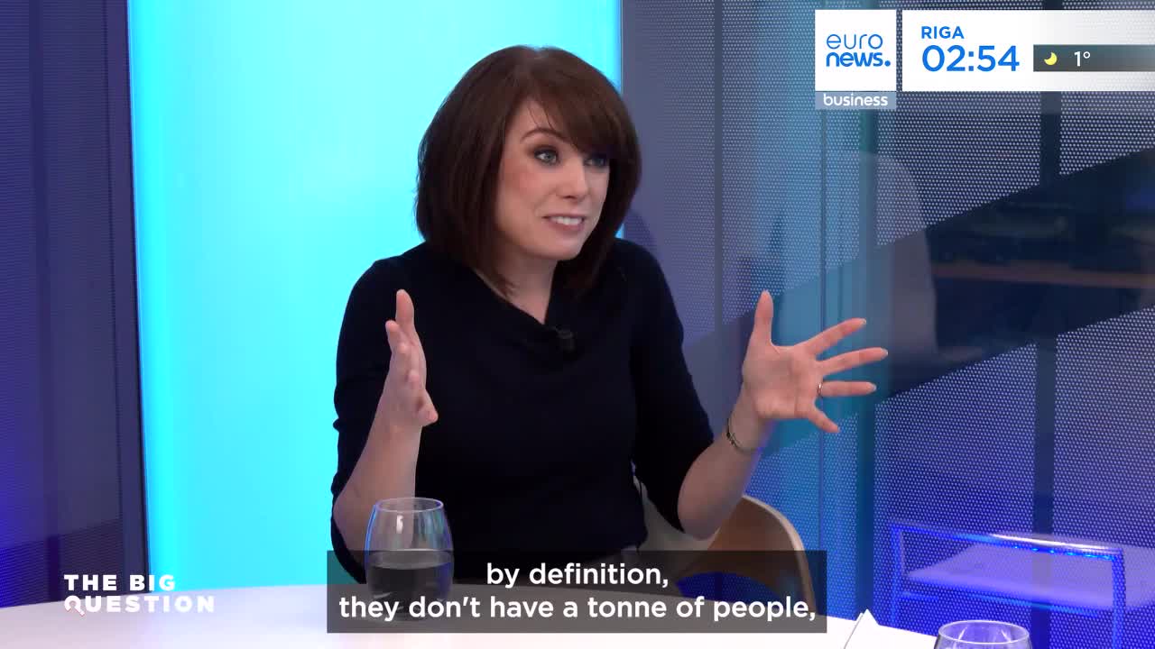 A woman with short brown hair is speaking animatedly, gesturing with both hands. A glass of water sits on the table in front of her, and a Euronews overlay indicates the broadcast is from Riga.
