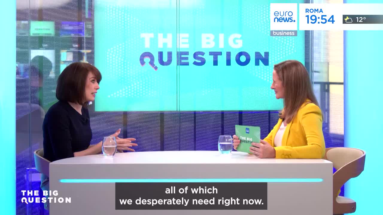 Two women sit at a table in a studio, the backdrop displaying "THE BIG QUESTION." The woman on the left, wearing a dark top, gestures with her hands as she speaks. The woman on the right, in a bright yellow blazer, holds a green booklet and listens intently.