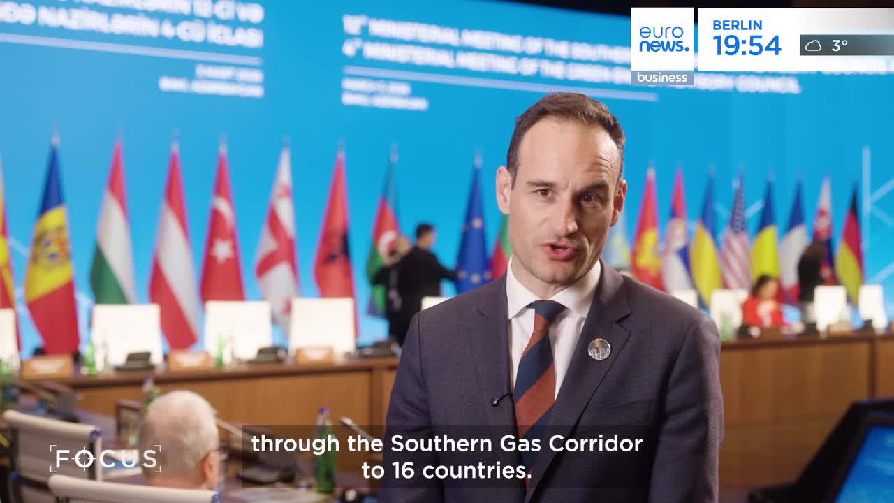 A man in a suit speaks directly to the camera, with flags of various nations displayed behind him. In the background, people are seated at a long table, and a few figures move between the rows of flags.