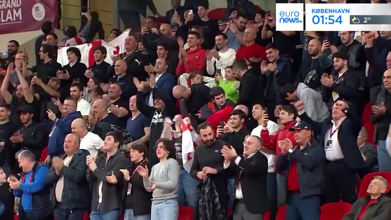 The crowd erupts in applause, many holding up phones to capture the moment. A man in a black suit and tie, wearing a lanyard, enthusiastically claps his hands.