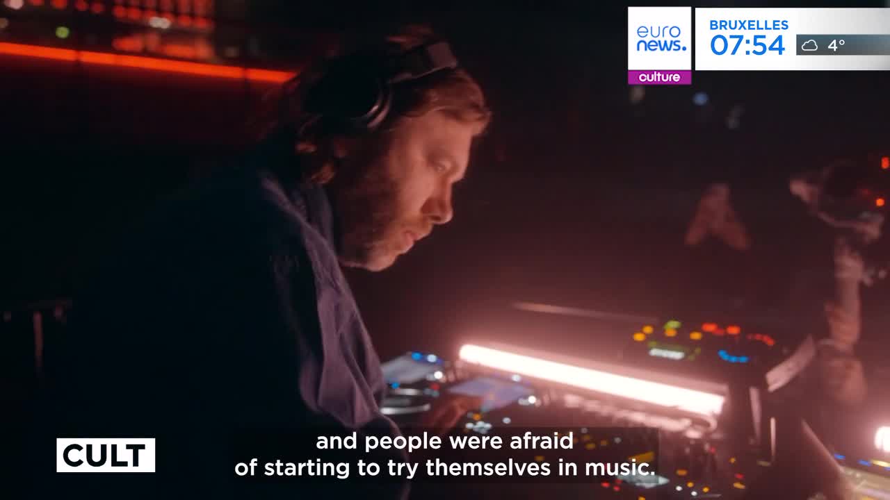 A man with headphones on leans over a DJ setup, his hands hovering above the controls. Red lights cast a warm glow over the equipment and his focused face.