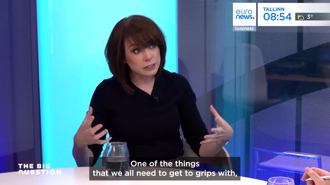 A woman with short brown hair gestures with both hands as she speaks. A glass of water sits on the table in front of her, and a graphic displays "THE BIG QUESTION" on the left.