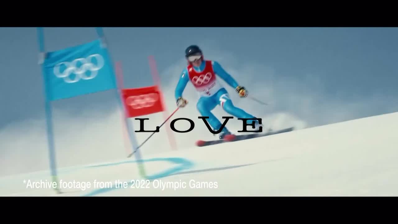 A skier in a bright blue suit carves down a snowy slope, poles extended. The Olympic rings fly on flags marking the course, a reminder of the competition in France.