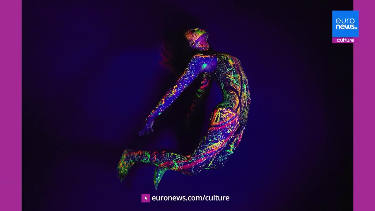 A figure contorts mid-air, limbs splayed, their body a canvas of vibrant neon paint under a blacklight. The fluorescent patterns pulse with electric blues, greens, and pinks against the deep purple backdrop, a striking visual from a performance in France.