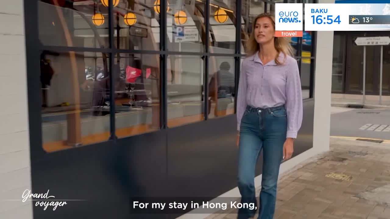 A woman in a light purple shirt and blue jeans walks past a building with large windows.  Reflected in the glass, I can see people inside and a glimpse of the street beyond.