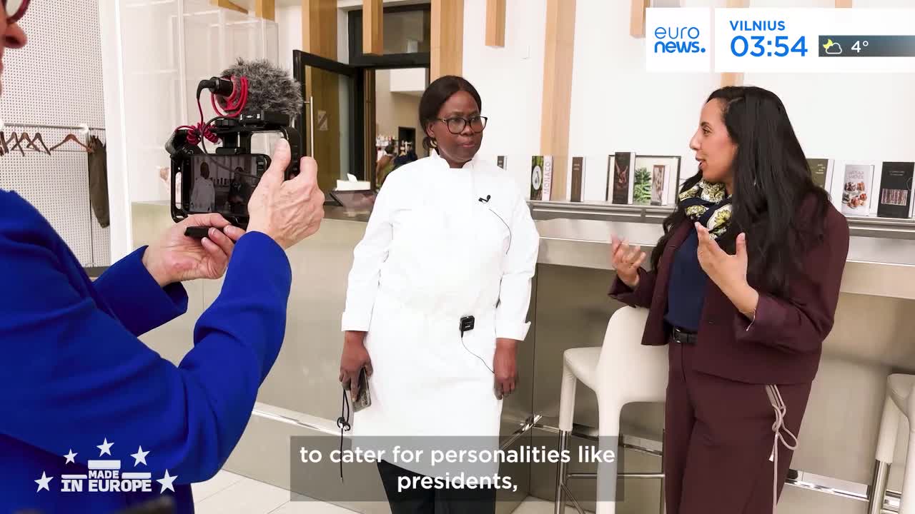 A person in a blue jacket films a woman in a chef's coat, who stands beside another woman speaking animatedly. The scene is set indoors, with shelves of what appear to be books or products behind the speakers.
