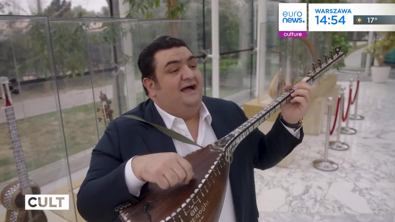 A man in a suit plays a long-necked stringed instrument, his mouth open as if singing. Behind him, other instruments are displayed in glass cases.