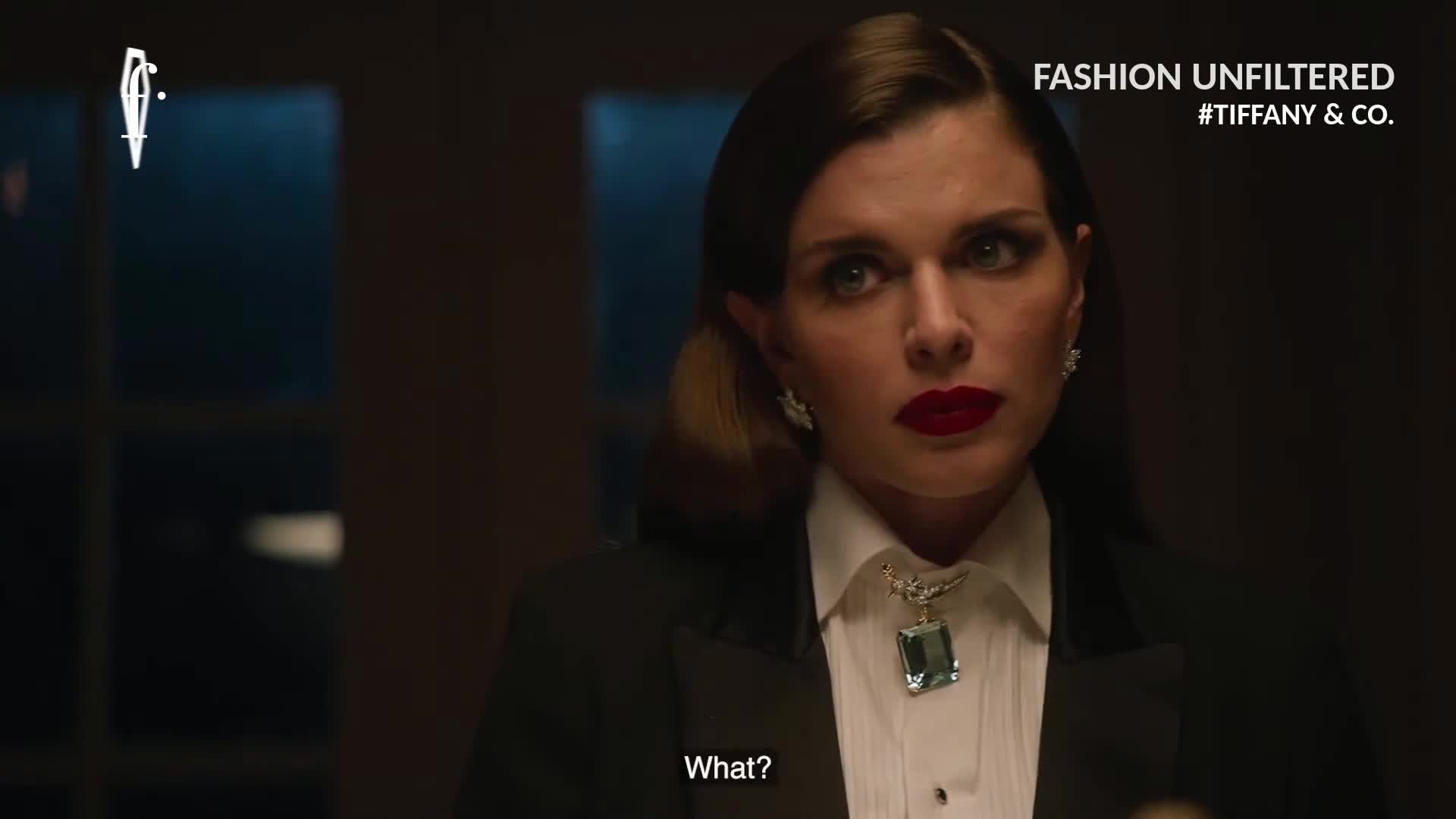 The woman in a tuxedo jacket and a striking necklace stares directly ahead, her red lips forming the word "What?" The scene is set in a dimly lit room, likely in France, with the FashionTV Europe logo visible in the corner.
