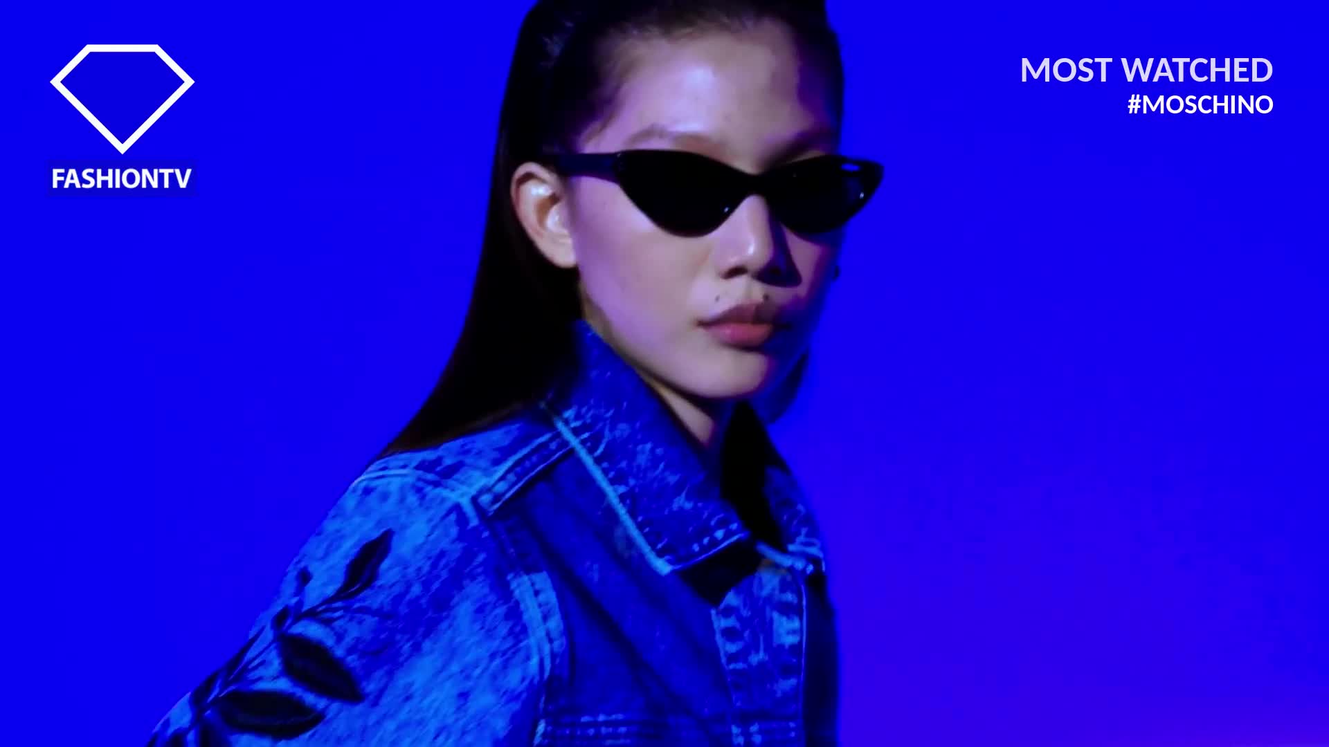 A model, wearing dark sunglasses and a denim jacket, faces the camera against a vibrant blue backdrop. The FashionTV logo is visible, and the hashtag #MOSCHINO indicates the featured designer.
