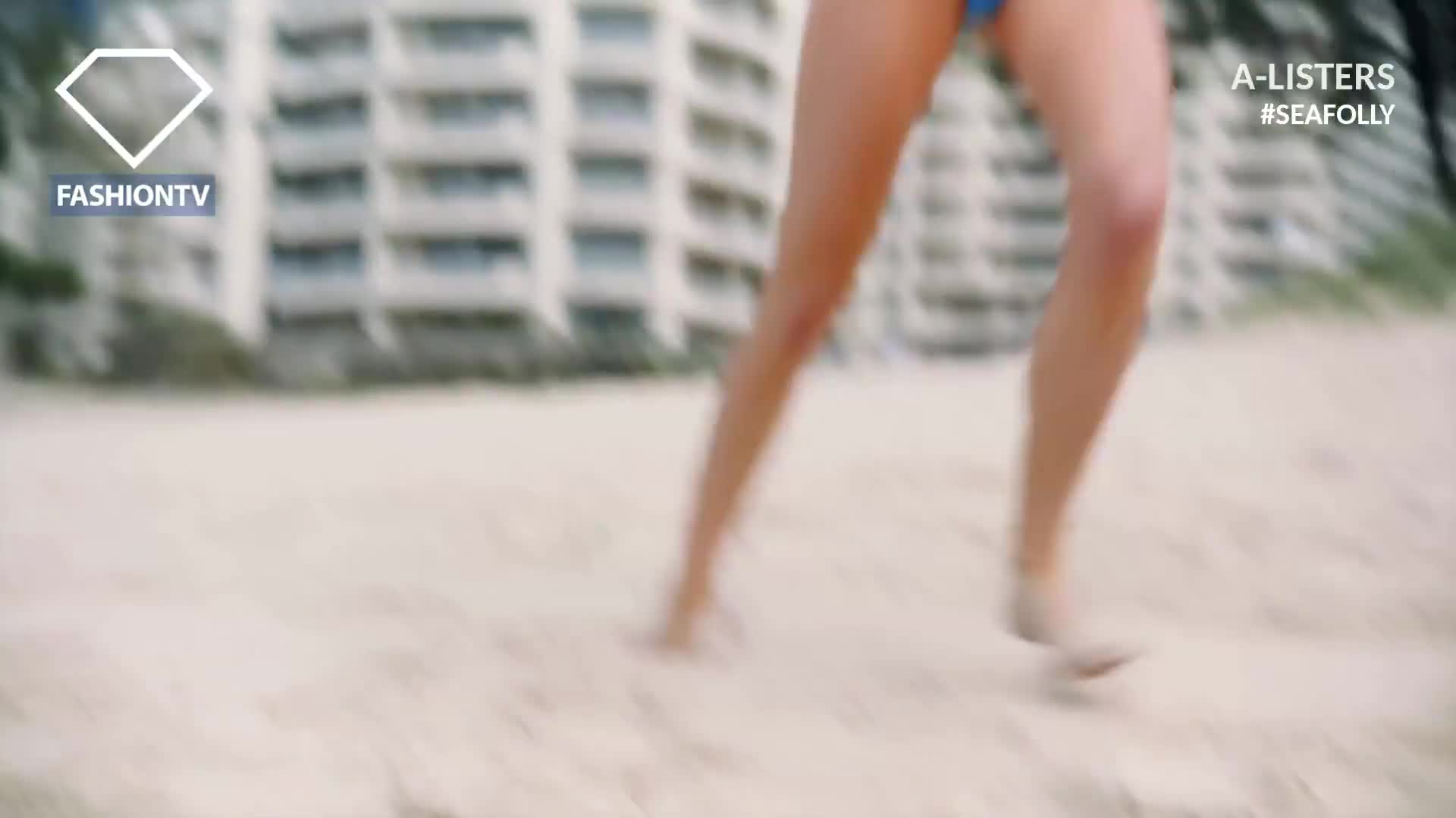 Bare legs in a blue swimsuit are sprinting across the sand, the feet kicking up small puffs of the beach. The FashionTV logo is in the corner, and the buildings in the background suggest a coastal location, perhaps France.
