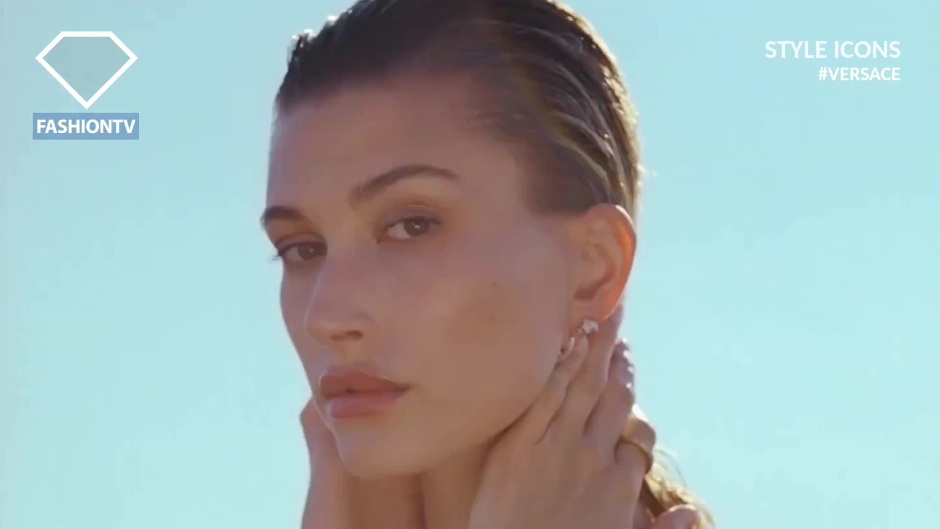 The model's hands gently cradle her neck as she looks toward the camera, her wet hair glistening in the sunlight. A FashionTV logo and the Versace hashtag appear on the screen, hinting at a shoot in France.
