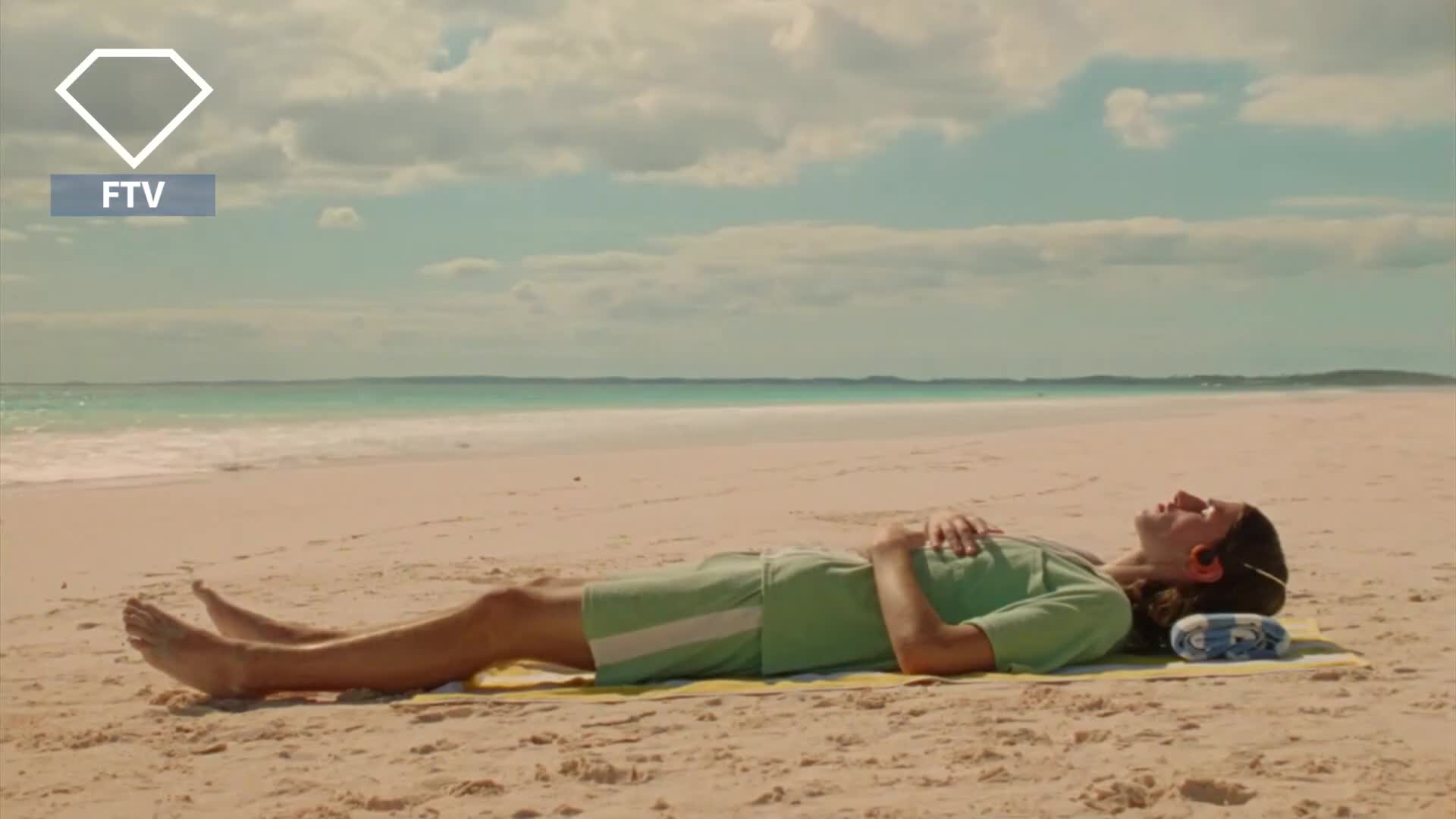 A person in a green outfit relaxes on a beach, eyes closed, a small pillow under their head. The ocean stretches out in the distance under a bright, cloudy sky.
