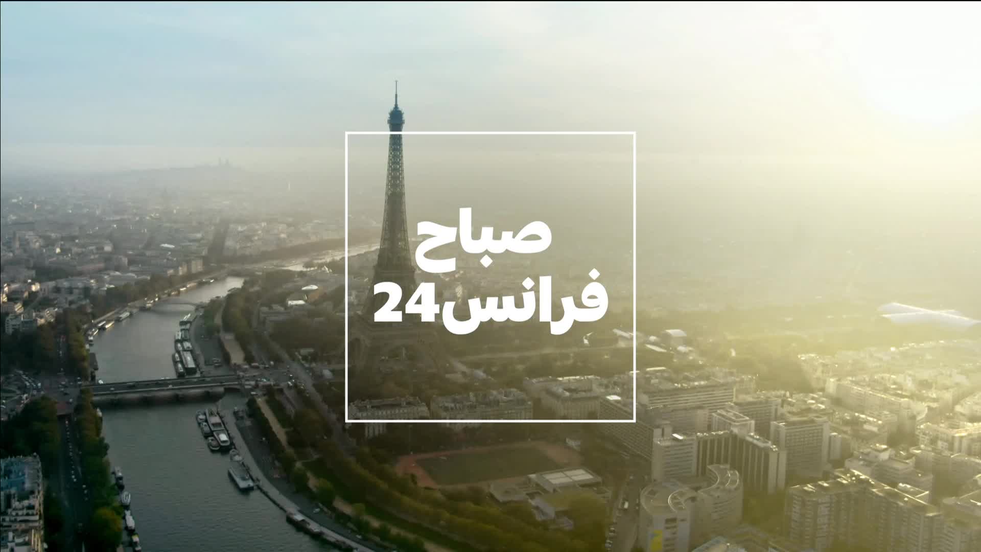 The Eiffel Tower pierces the morning haze over Paris, as the France 24 logo, written in Arabic, frames the scene. The Seine River snakes through the city, lined with buildings and boats.
