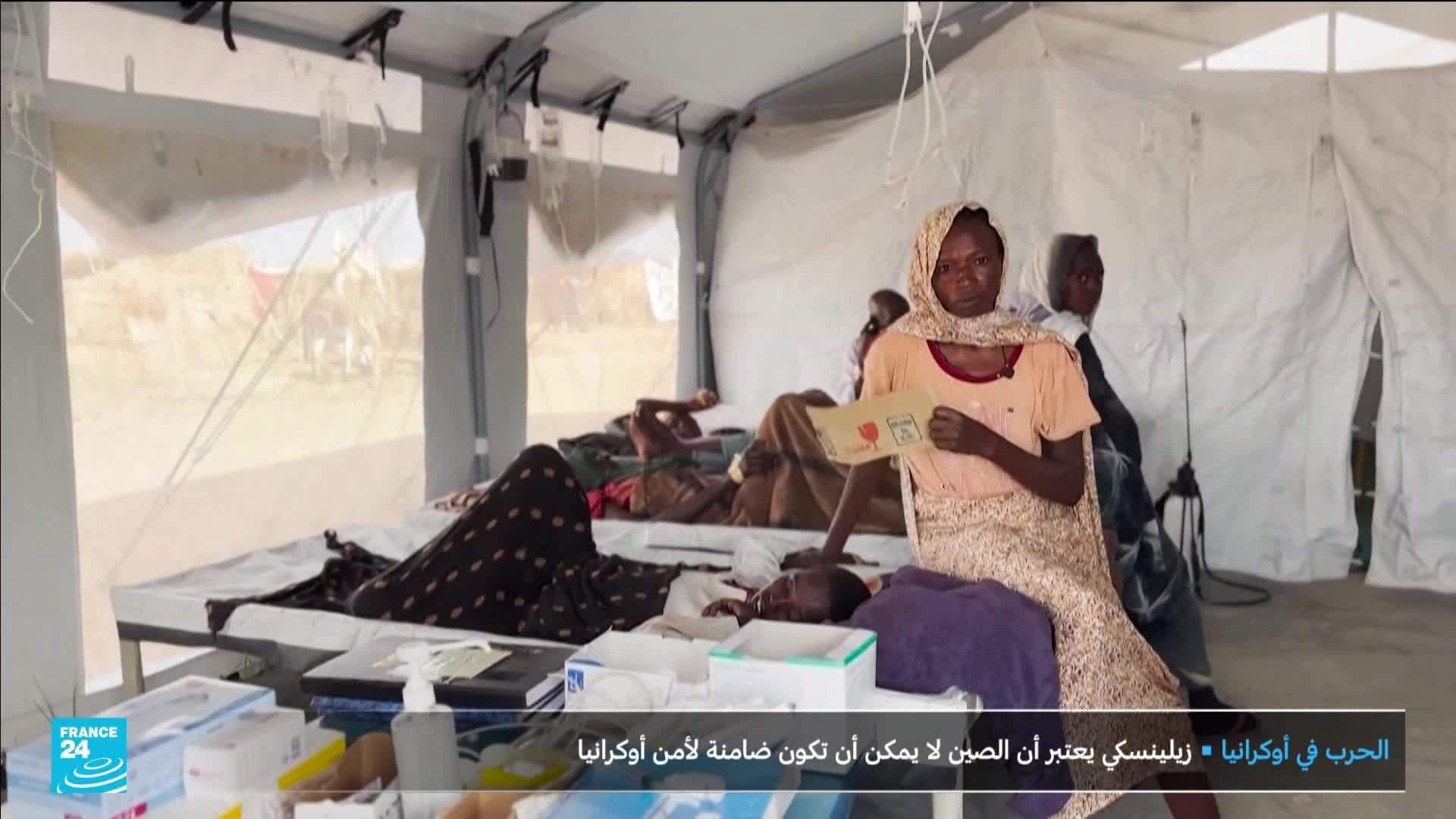 Inside a tent, several people rest on beds, some hooked up to IV drips. A woman in a headscarf holds a small card, while a France 24 logo appears in the corner.

