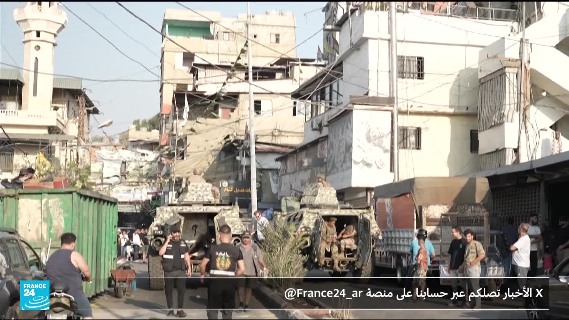 Two military vehicles, camouflaged and bulky, are parked on the narrow street, soldiers visible inside. People mill about, some watching, others going about their business amidst the tension.

