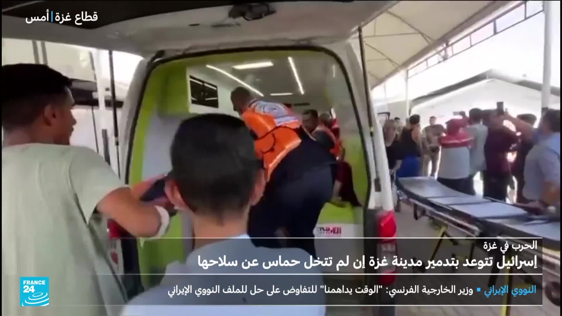 Men in orange vests are helping someone into the back of an ambulance. A crowd gathers nearby, and a stretcher sits ready on the ground.
