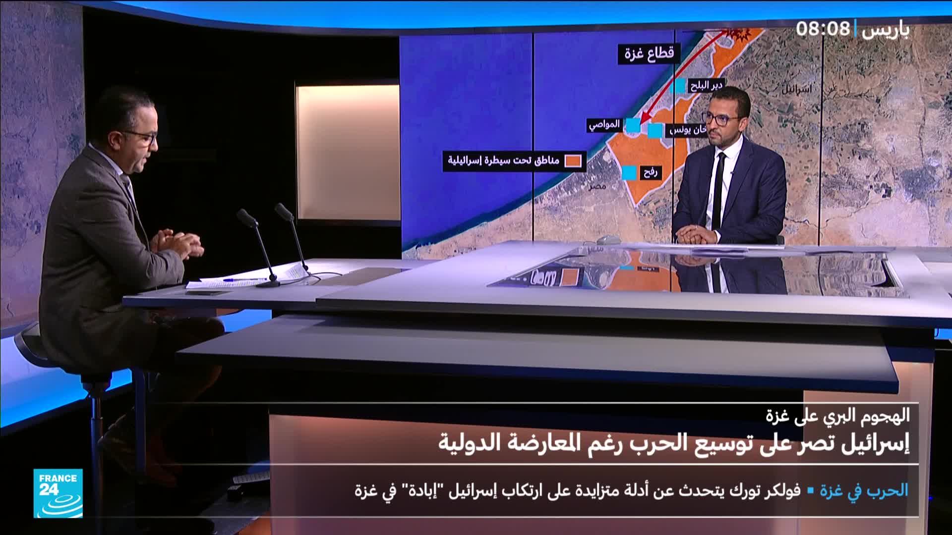 Two men in suits sit at a desk, facing each other, with microphones between them. Behind them, a map of Gaza is displayed on a screen with Arabic text and the France 24 logo in the corner.
