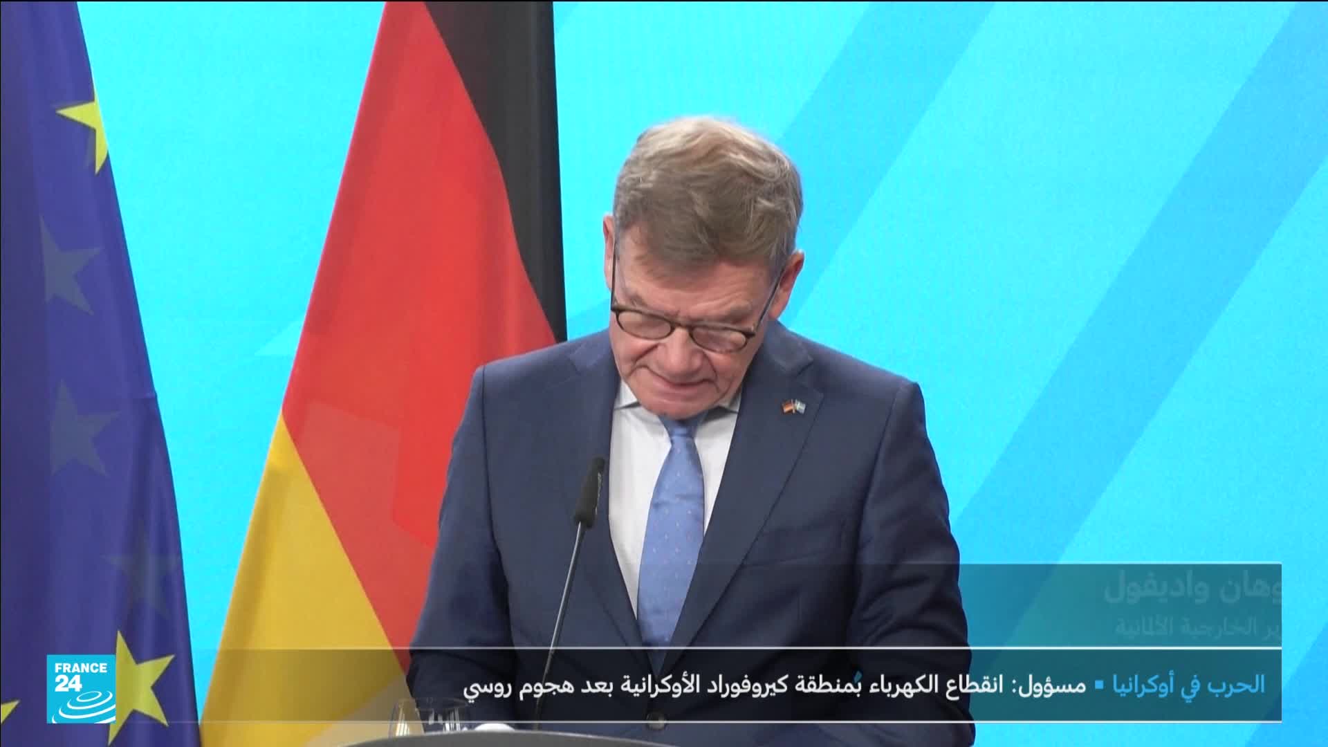 A man in a blue suit, glasses perched on his nose, reads from a document. The backdrop includes the German flag and the France 24 logo, with Arabic text scrolling at the bottom.
