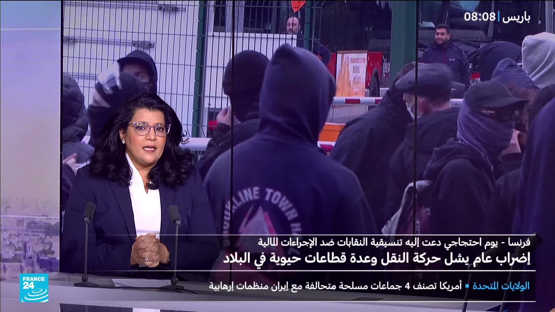 A news anchor in France is reporting on a protest, with a crowd of people in dark clothing and face coverings visible behind her. The backdrop shows a metal fence and a few figures watching the scene.
