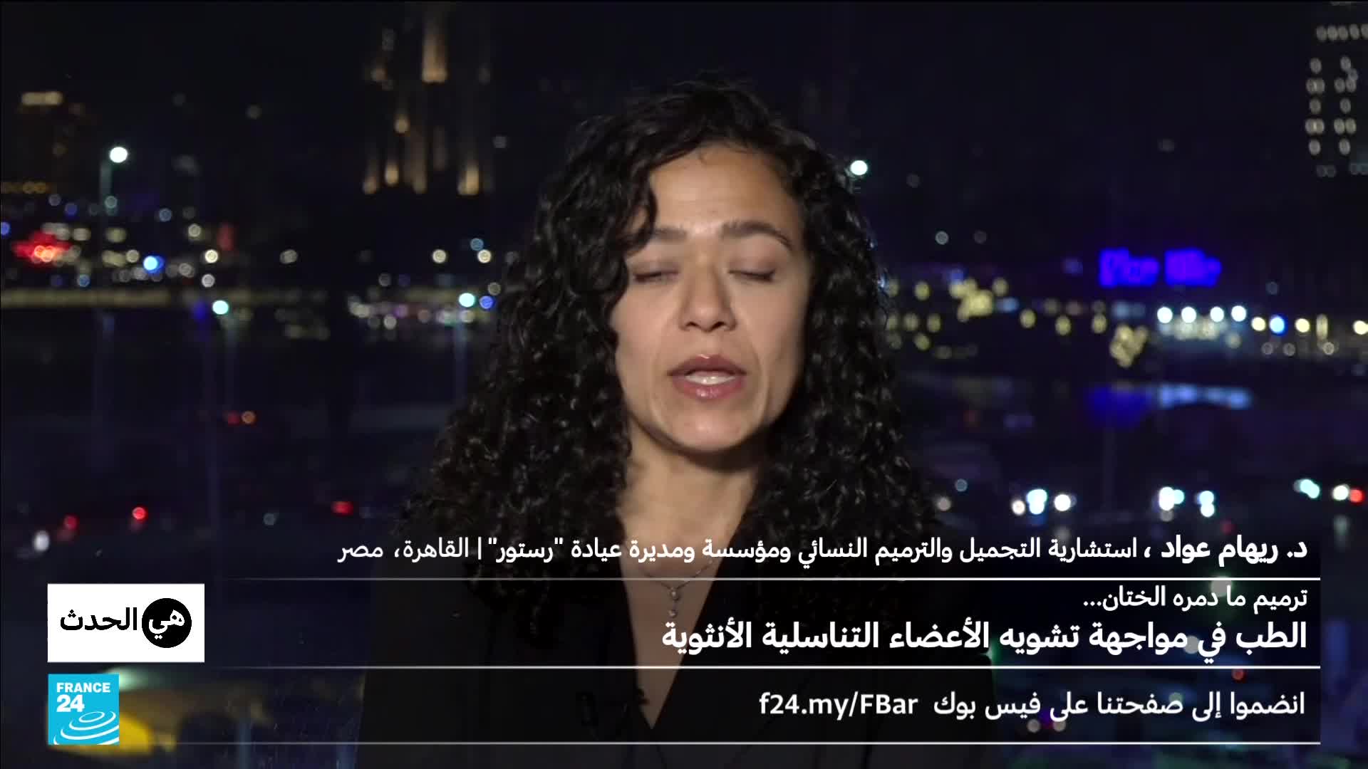 A woman with dark, curly hair speaks directly to the camera. Behind her, the lights of a city at night twinkle across a body of water.