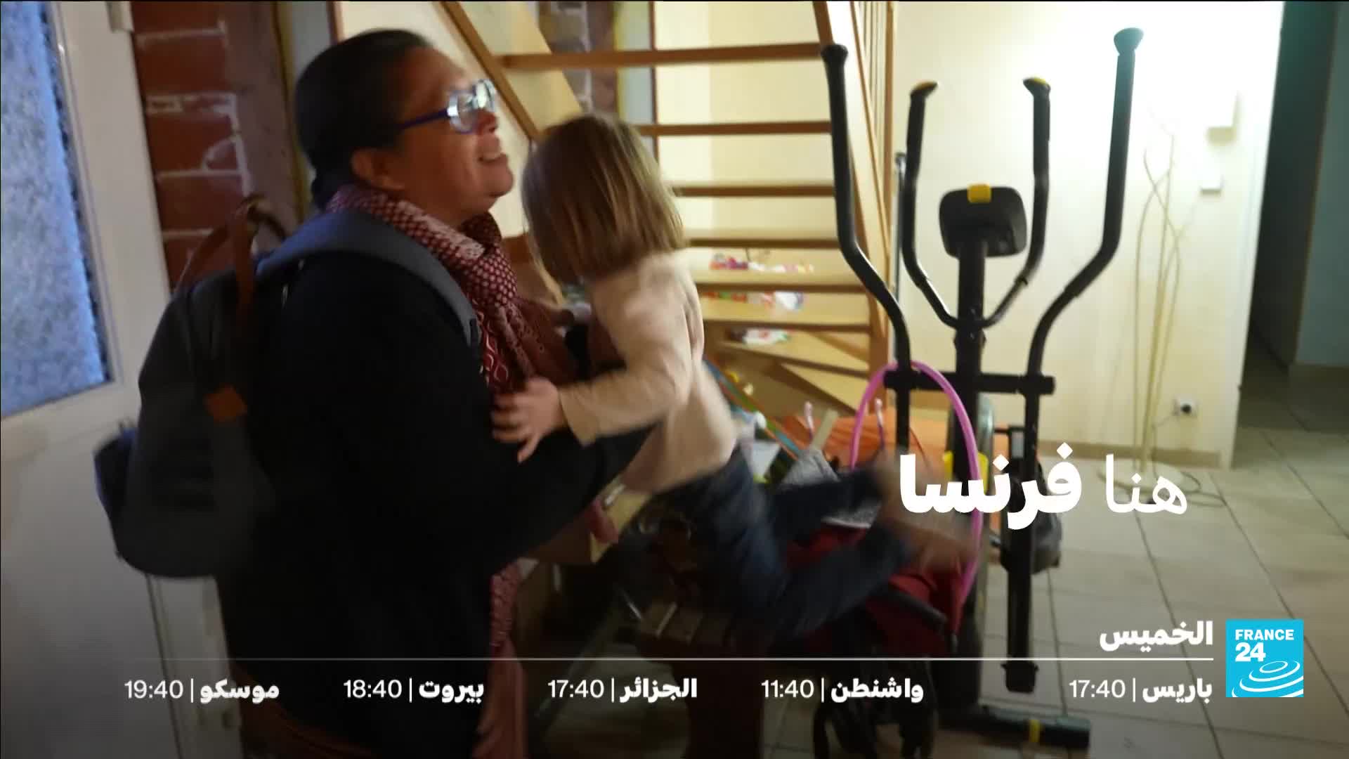 A woman with glasses and a patterned scarf embraces a small child in her arms. The child's legs are extended, and their body is angled as if being swung.