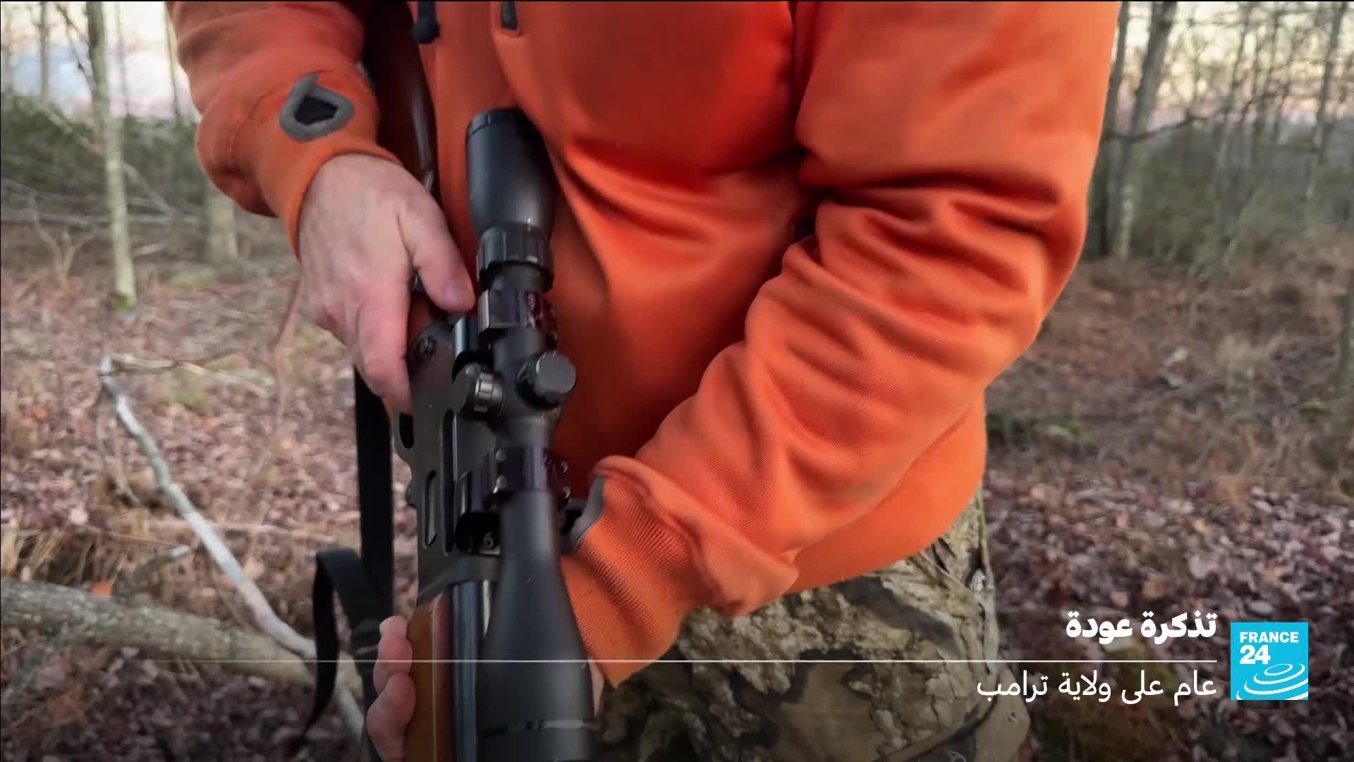 A person in an orange jacket is holding a rifle with a scope. The woods behind them are bare, with fallen leaves on the ground.