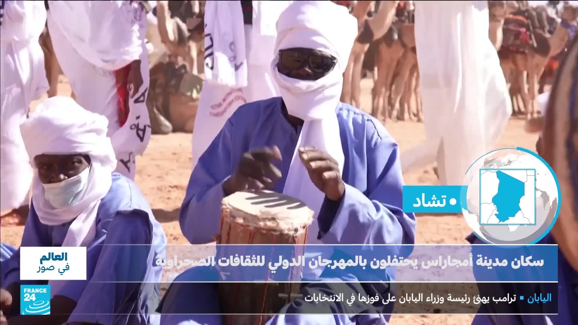 A man in a blue robe and white headwrap plays a drum, his hands a blur against the taut skin. Behind him, others in similar attire gather, some with camels in the distance, all under a bright, dusty sky.