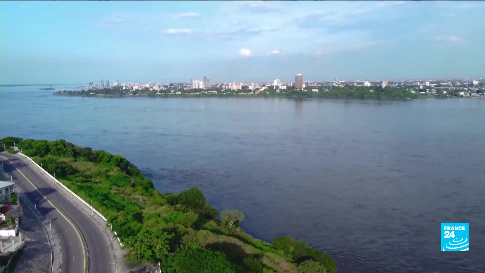 A wide river flows towards a distant cityscape under a bright sky. A paved road curves along a lush, green embankment in the foreground.