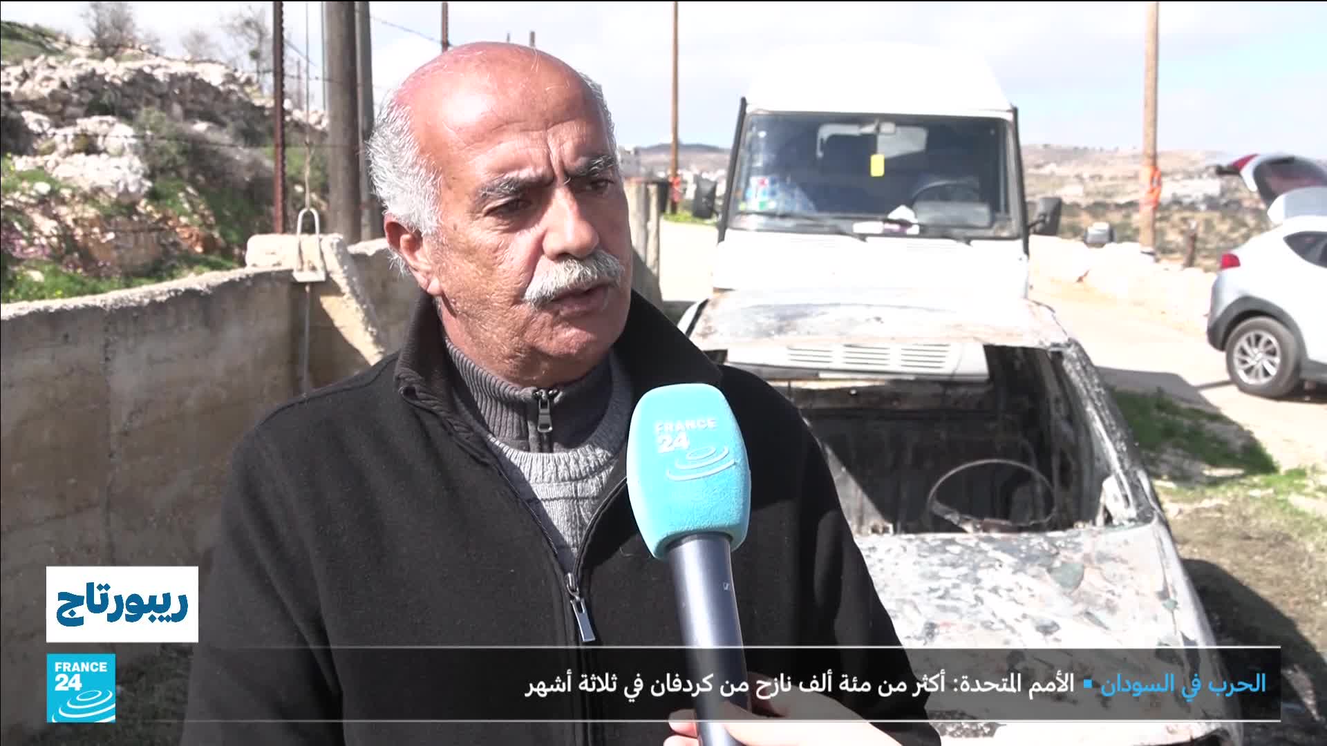 A man with a prominent mustache speaks into a France 24 microphone, his expression serious. Behind him, a white vehicle, its hood and interior scorched, sits on a dirt road.