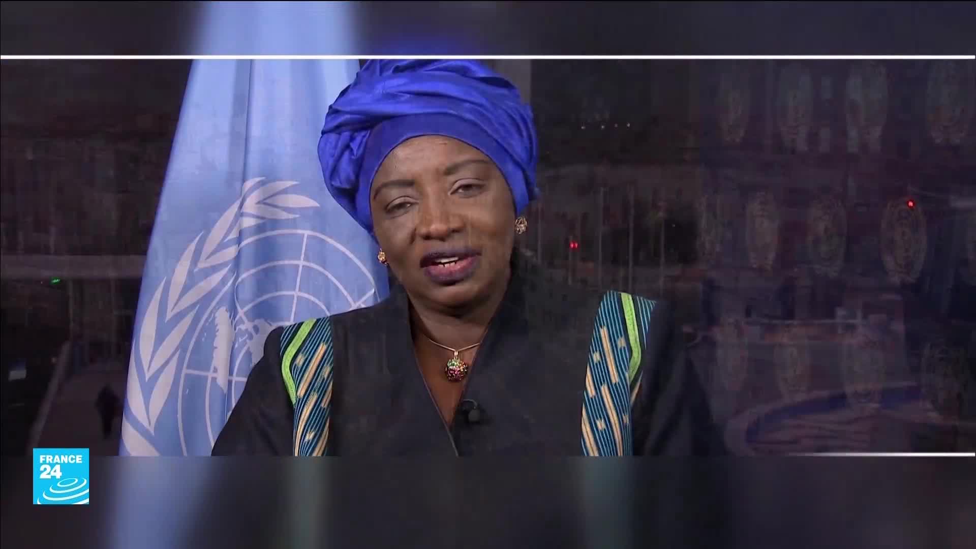A woman in a vibrant blue headscarf speaks, her dark jacket adorned with colorful, striped trim. Behind her, the United Nations flag hangs, and a reflection of city lights shimmers.