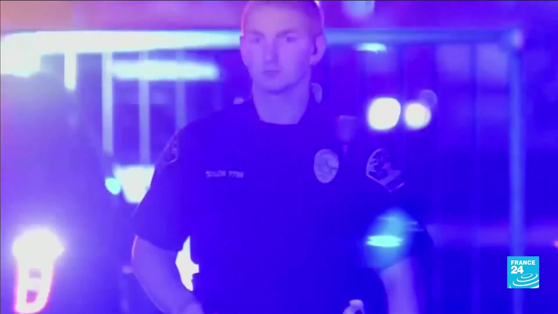 A young police officer stands in the blue and purple glow of flashing lights. He's looking forward, his uniform dark against the vibrant background.