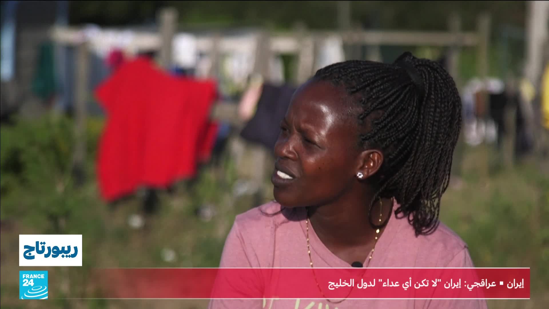 A woman with braided hair speaks, her expression earnest. Behind her, laundry hangs on a line, a bright red shirt catching the light.