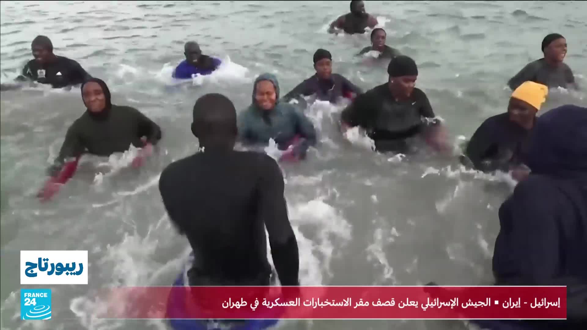 A group of people are wading through choppy water, some wearing dark hoodies and others bright yellow headwear. The waves splash around their chests as they move forward.