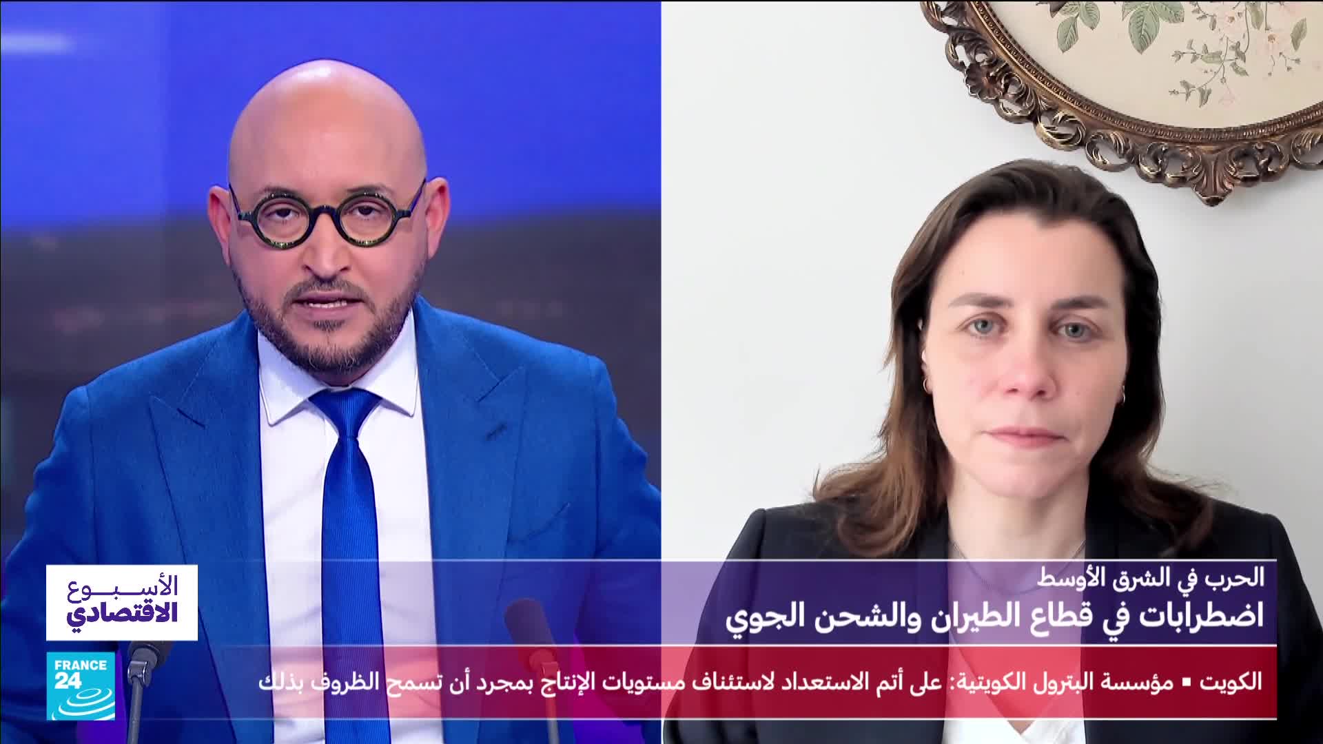 A man in a blue suit and tie speaks directly to the camera, his expression serious. Across from him, a woman in a dark blazer looks thoughtfully ahead, framed by an ornate mirror.