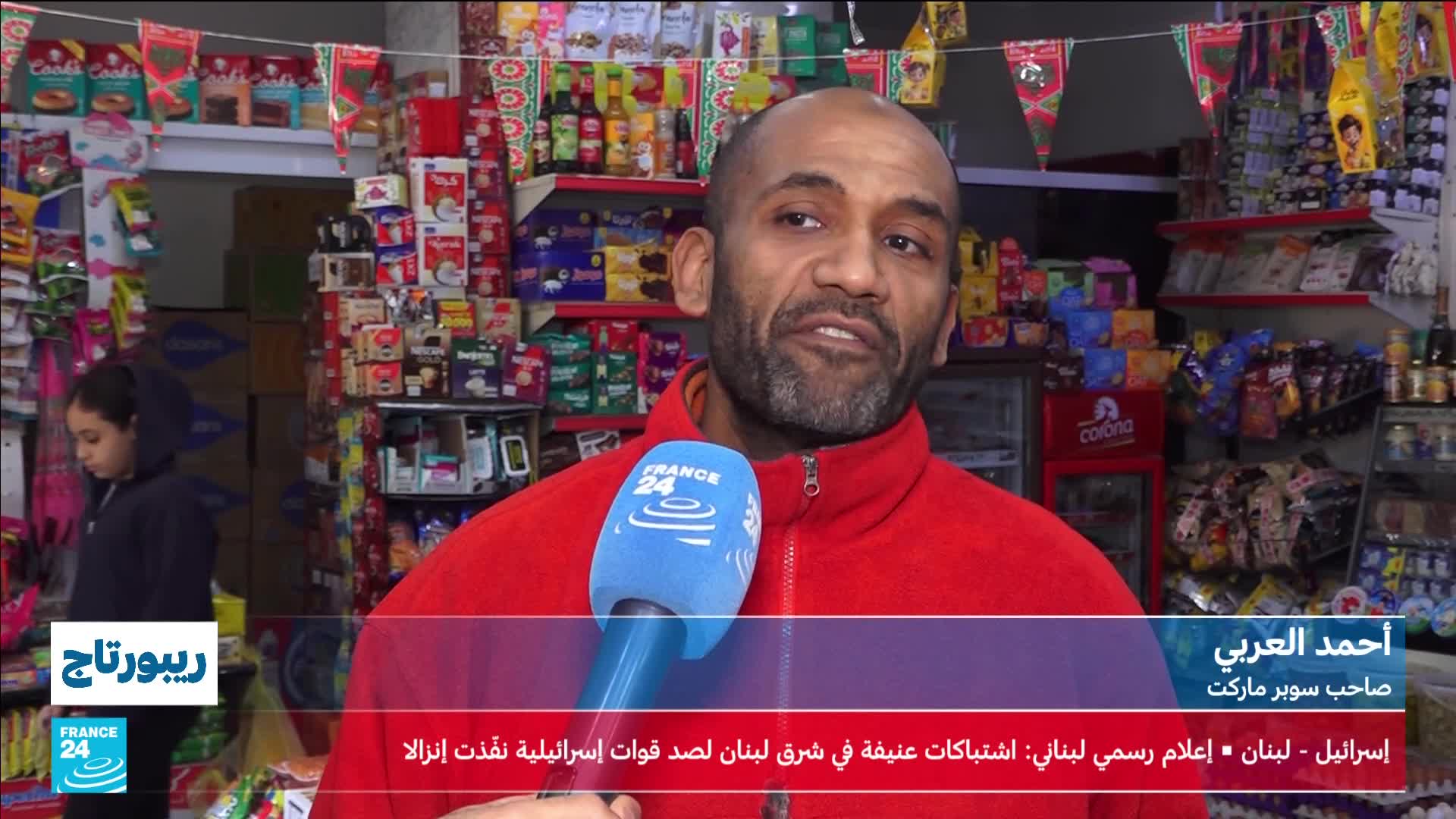 A man in a red fleece jacket speaks into a France 24 microphone. Behind him, shelves overflow with packaged goods in a small shop.