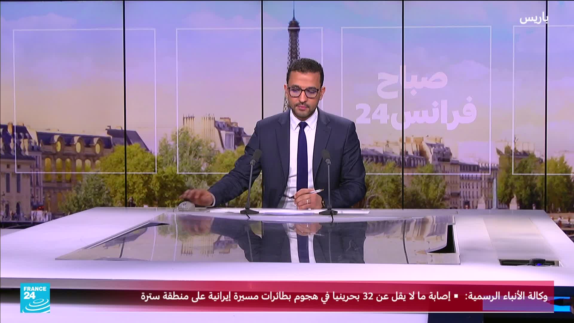 A man in a suit sits at a desk, holding a pen, with the Eiffel Tower visible in the background. A chyron at the bottom of the screen displays news from France 24.