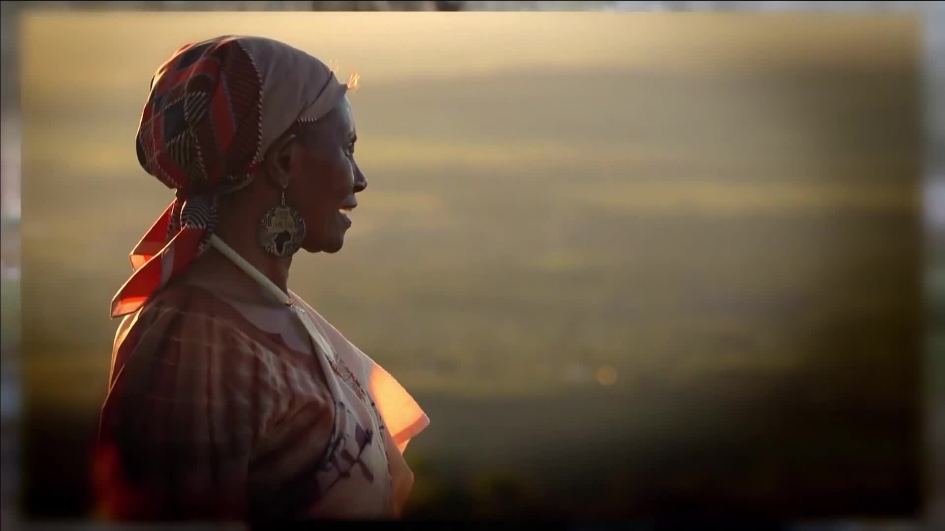 The sun catches the patterned headscarf and earrings of a woman as she looks out over a hazy, golden landscape. Her patterned dress is illuminated from the side, suggesting a warm, late afternoon light. The sun catches the patterned headscarf and earrings of a woman as she looks out over a hazy, golden landscape. Her patterned dress is illuminated from the side, suggesting a warm, late afternoon light.