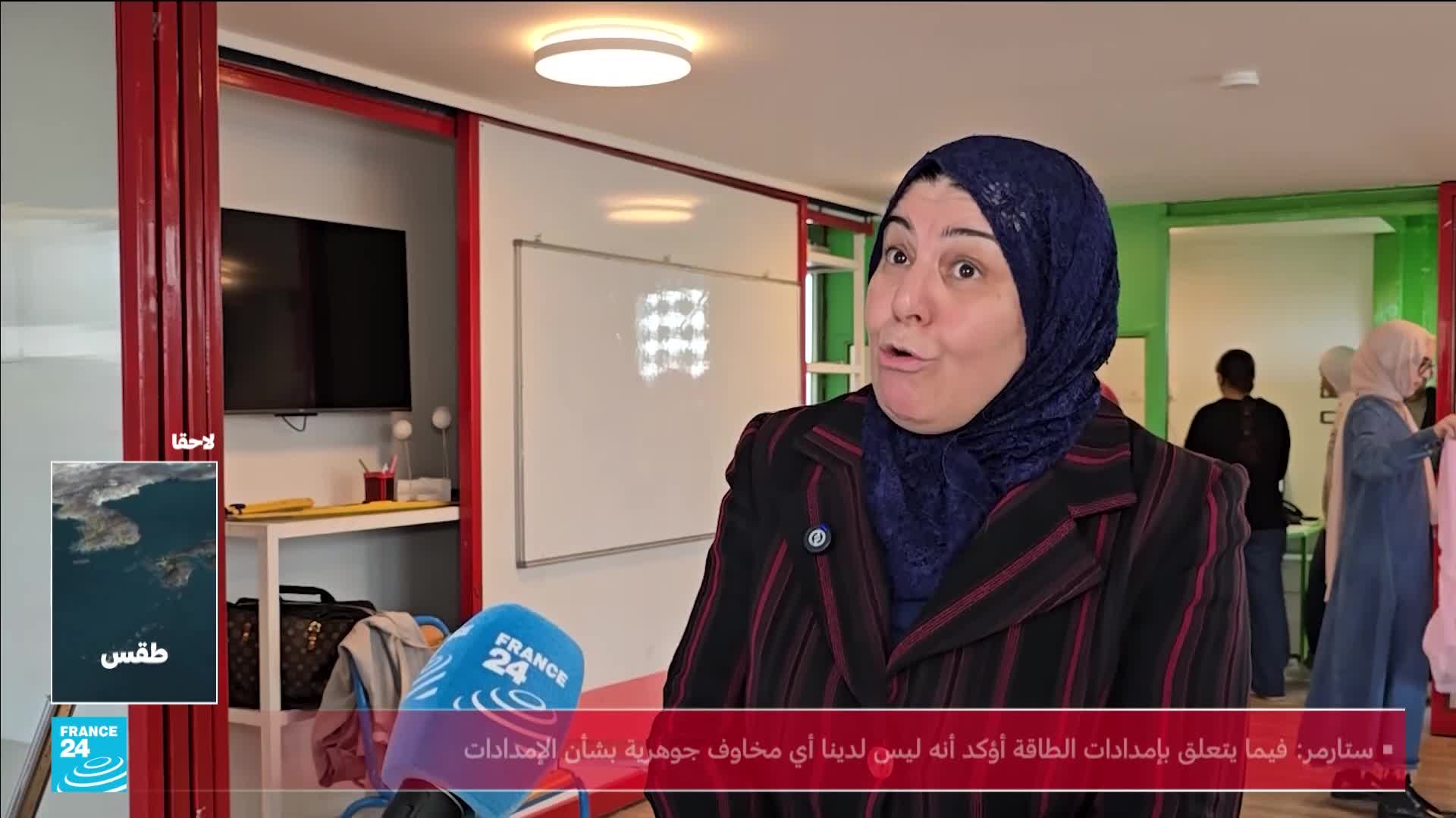 A woman in a hijab speaks animatedly into a microphone. In the background, other women are arranging fabric. A woman in a hijab speaks animatedly into a microphone. In the background, other women are arranging fabric.